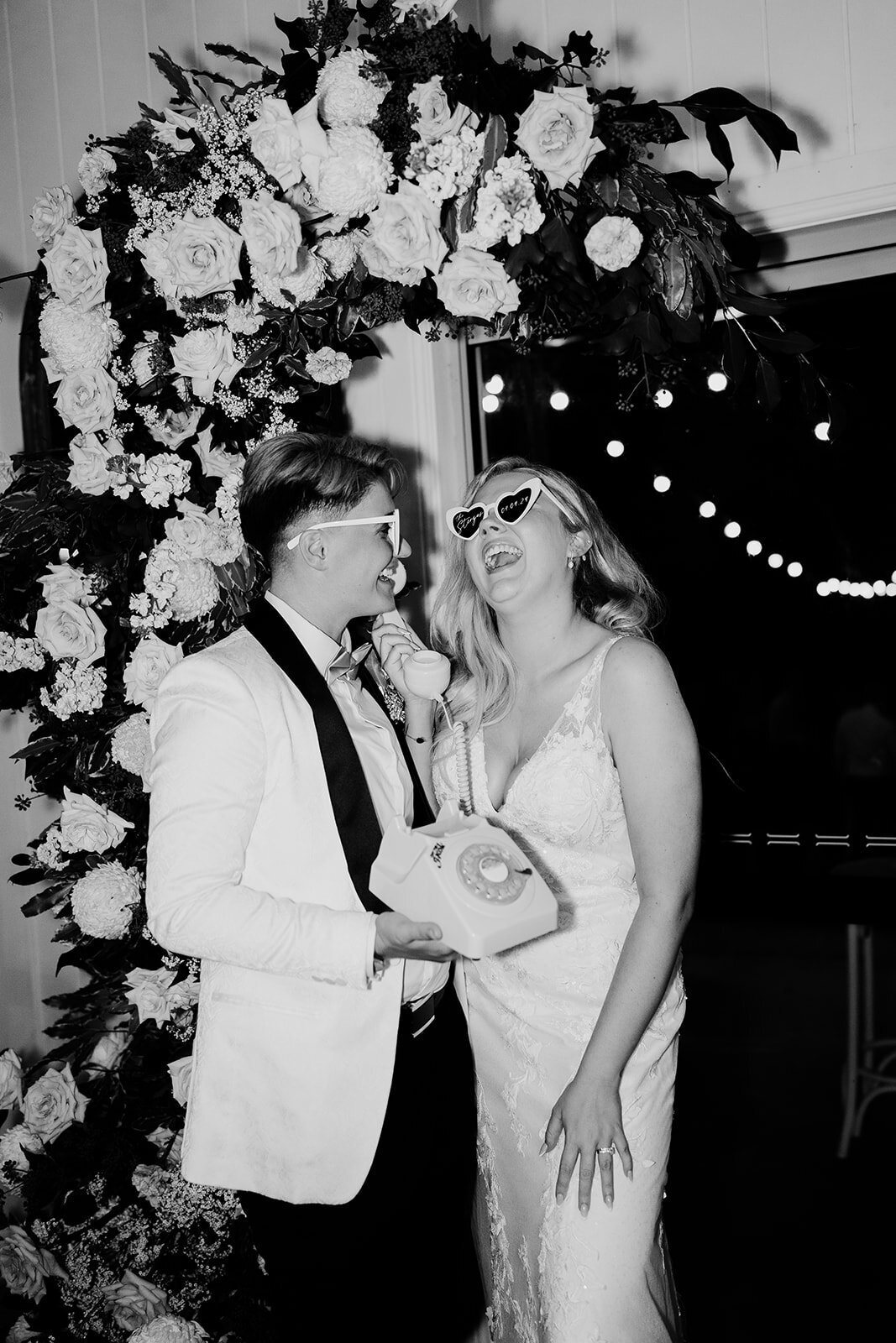 Black and white: a couple in novelty sunglasses laughing under a flower arch