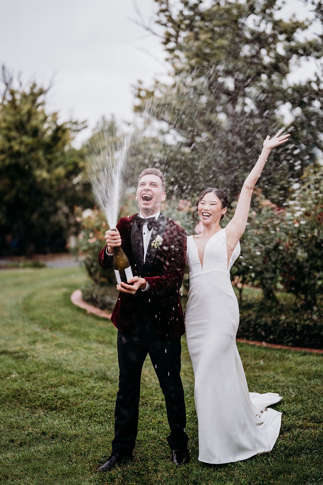 A groom spraying champagne as his bride throws her arm in the air on a garden lawn