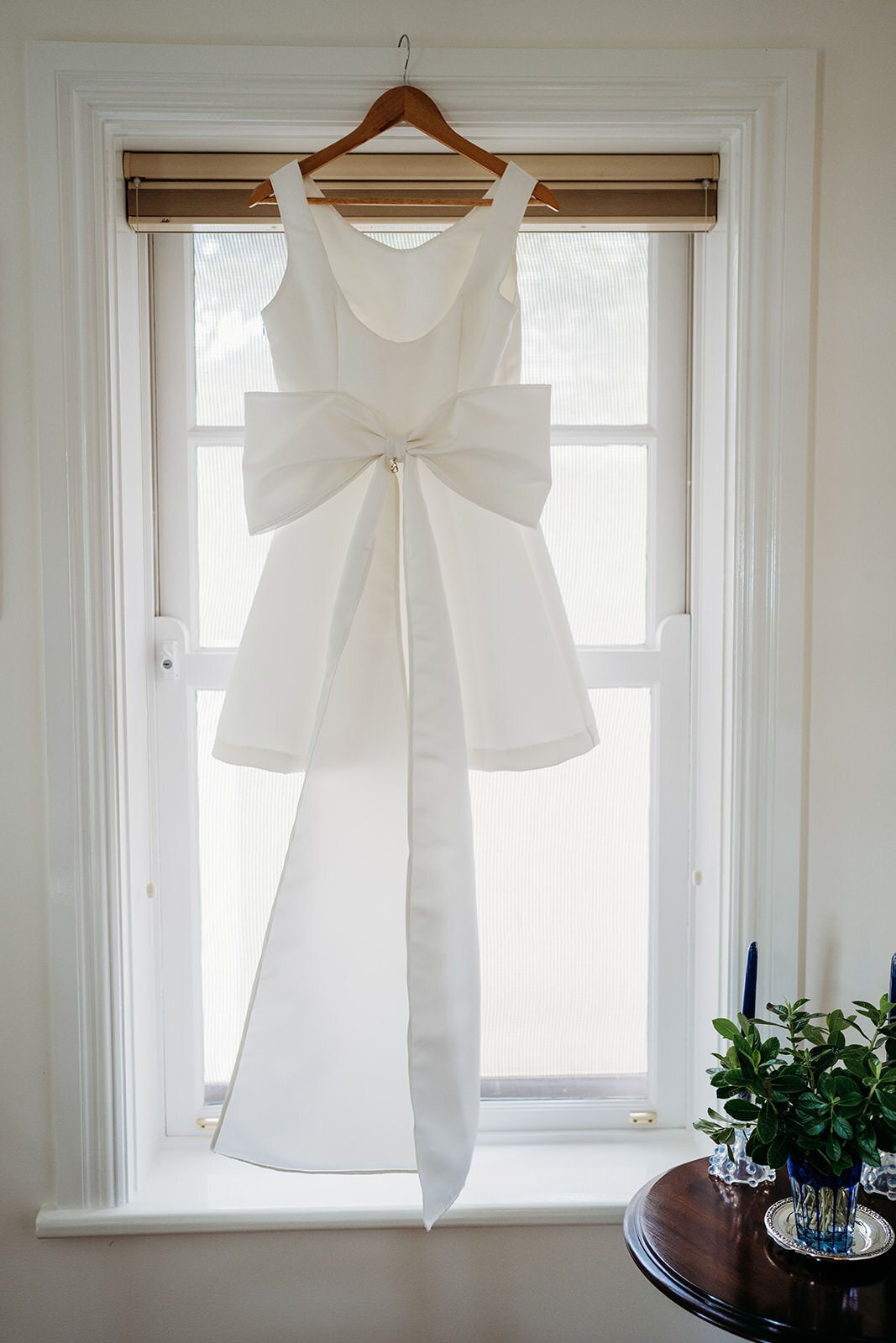 A wedding dress with a large bow hanging in a sunlit window