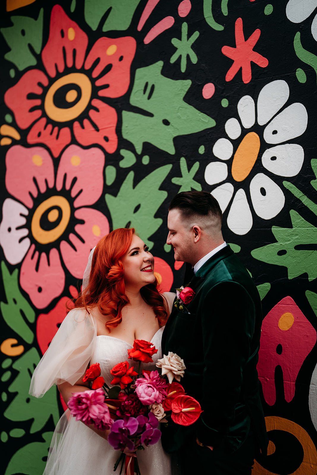 A bride with red hair and her groom in front of a bright flower mural