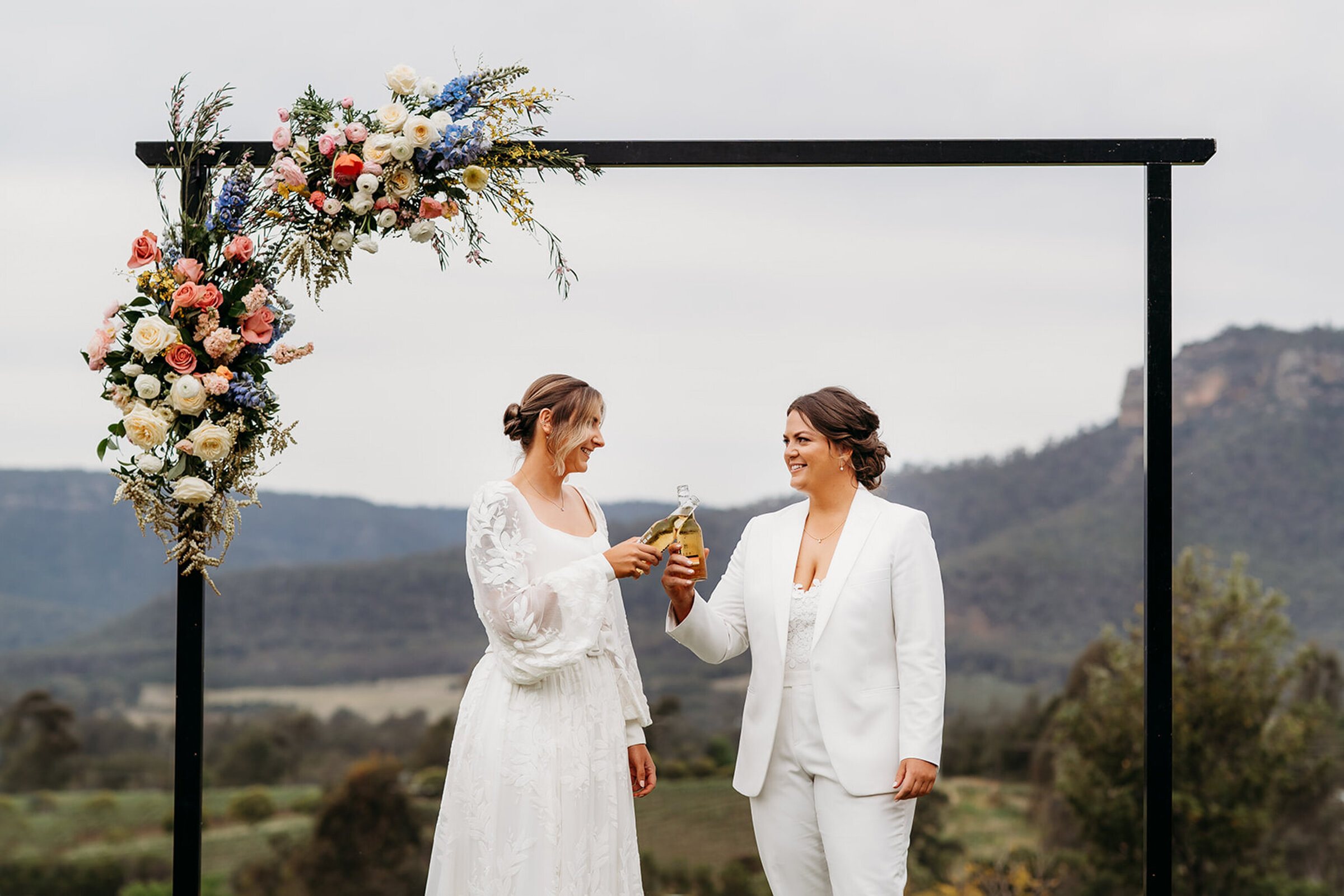 Two brides laughing under a flower arch with mountains behind them