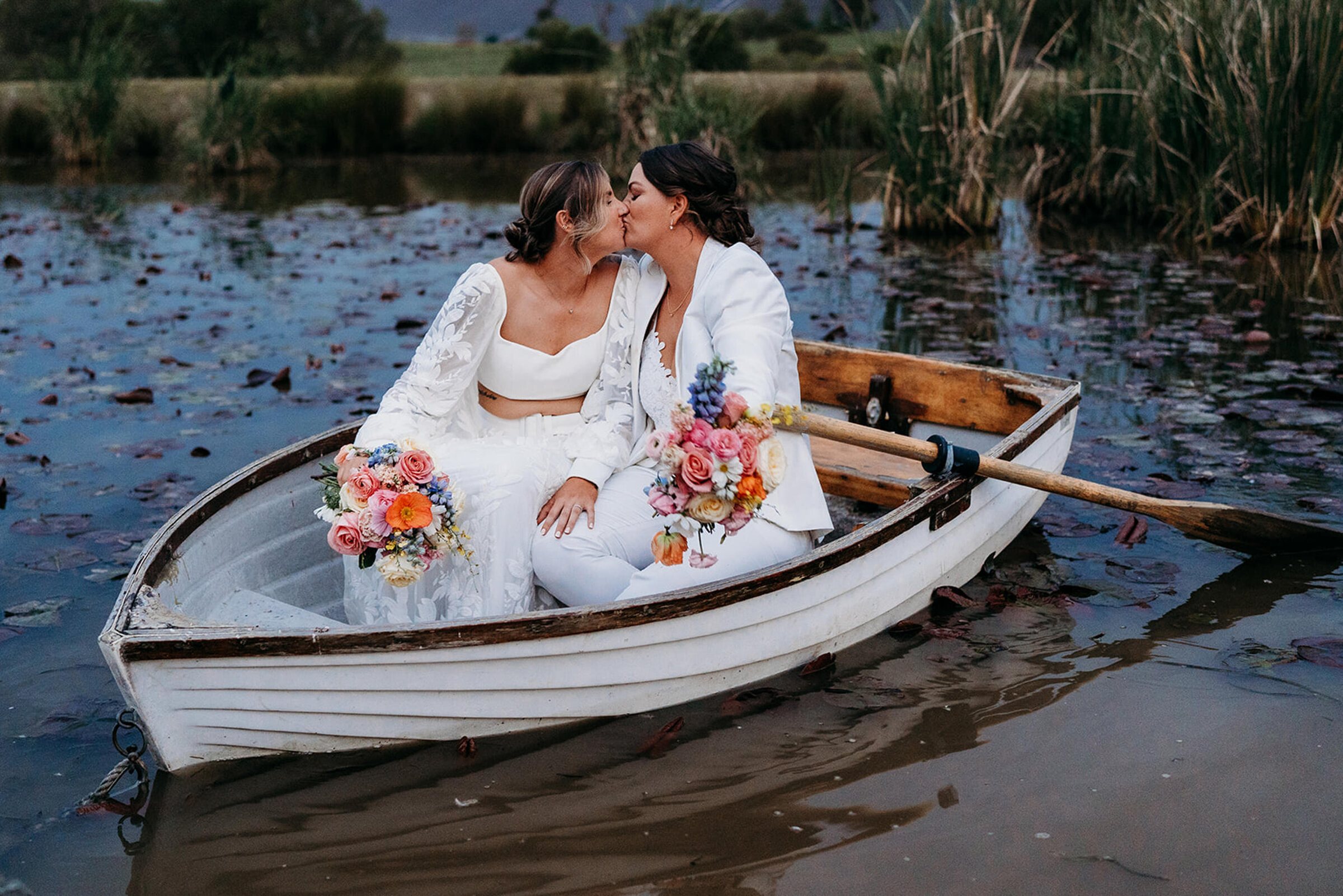 Two brides sharing a kiss in a small white rowboat on a lily pond