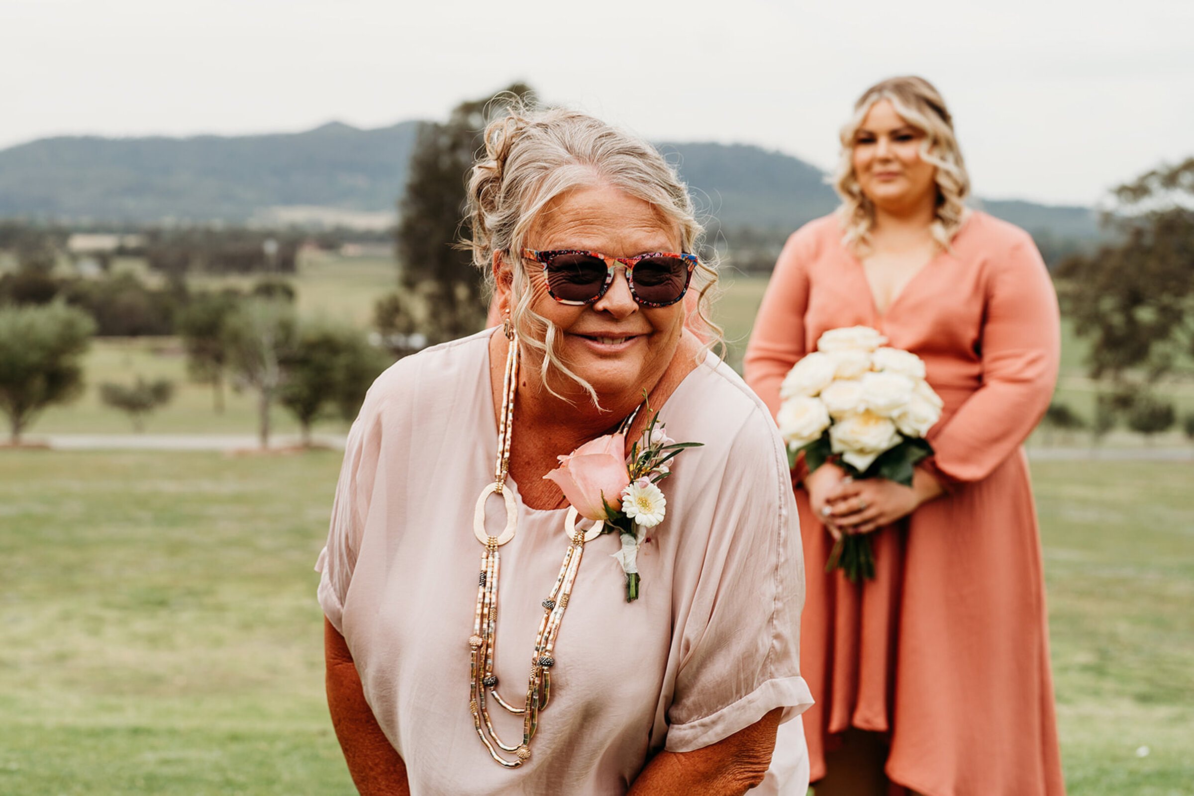 A wedding guest laughing in sunglasses beside a bridesmaid holding a bouquet