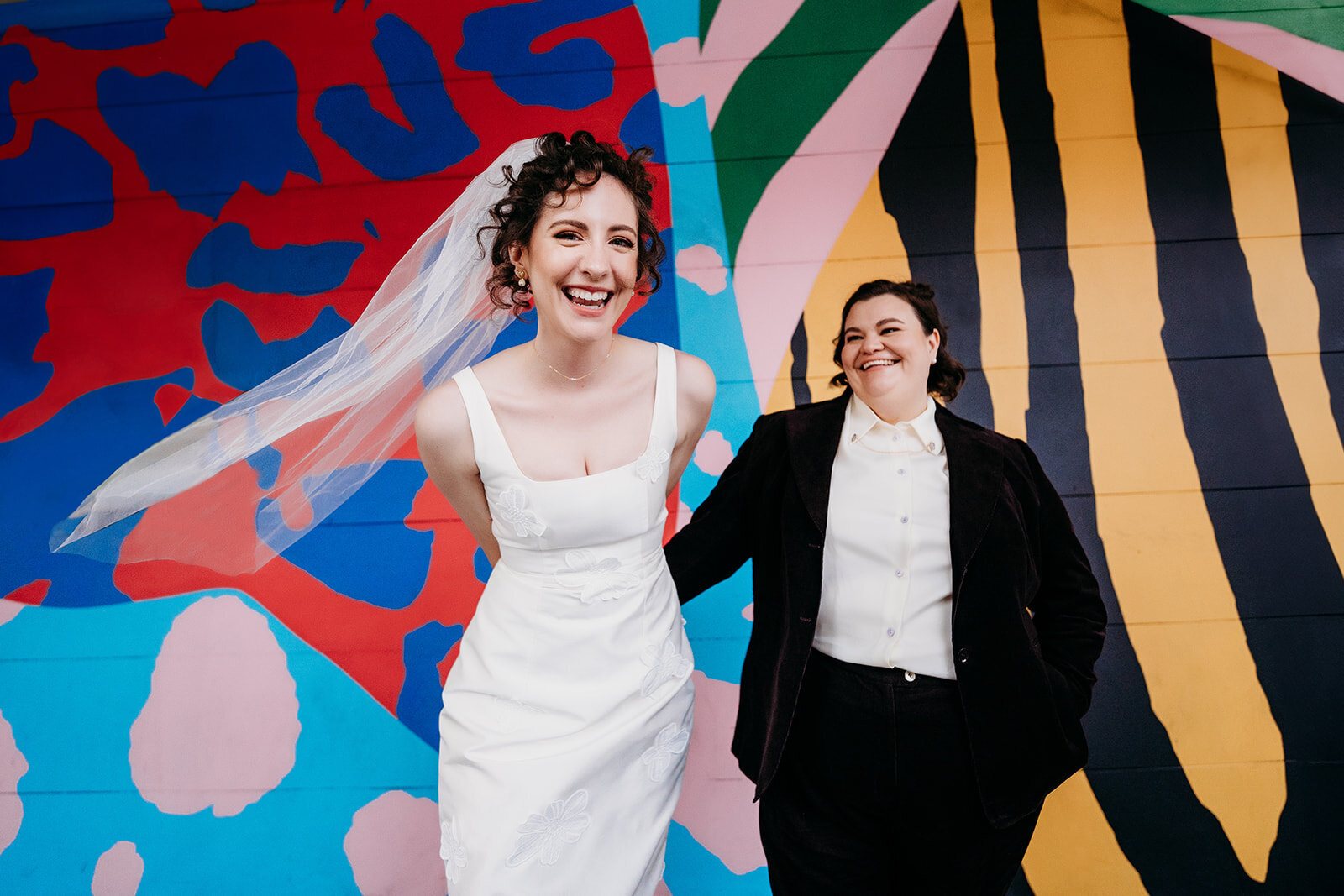 Two brides laughing in front of a colourful painted mural