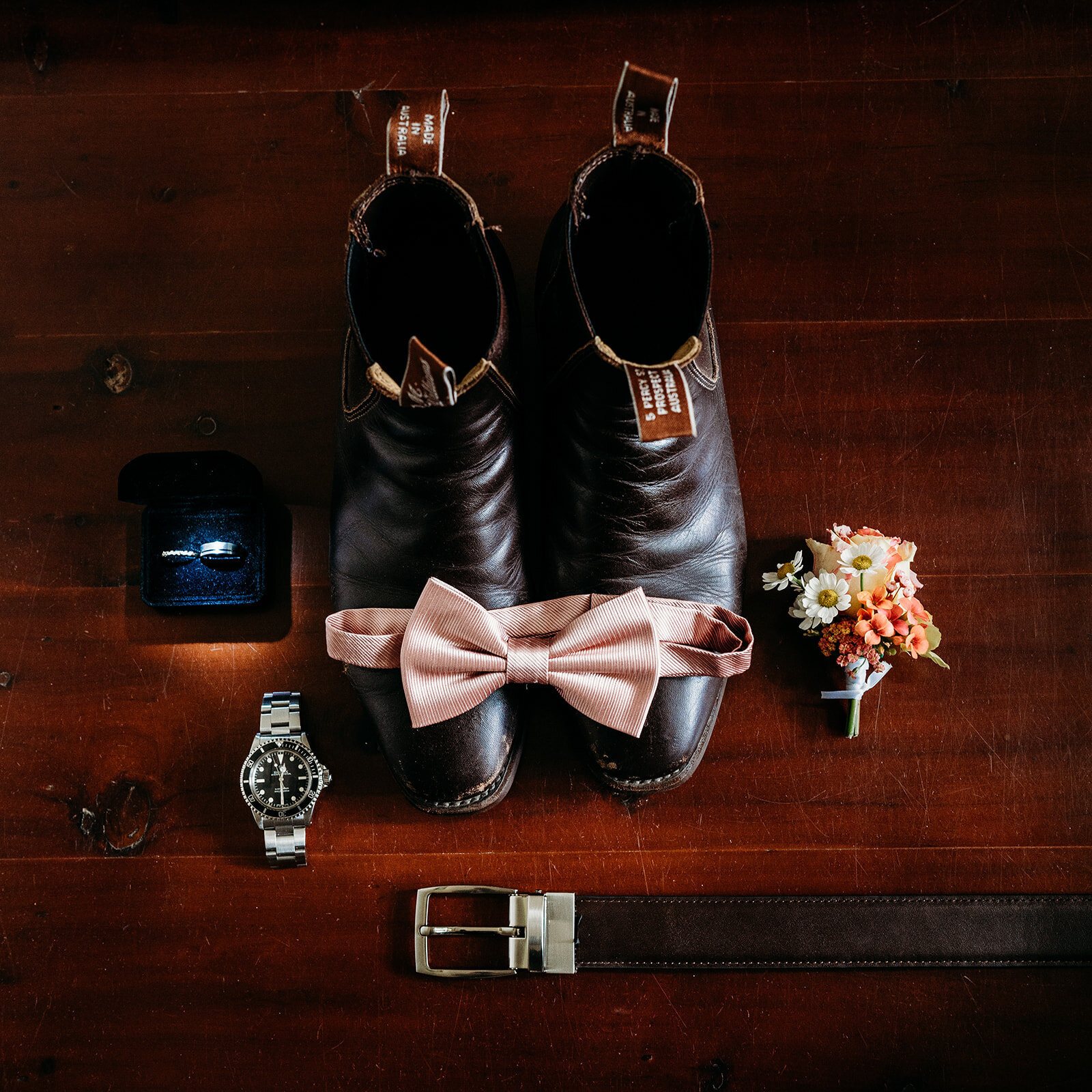 Black boots, a pink bow tie, a watch, a belt and a ring box laid out on timber