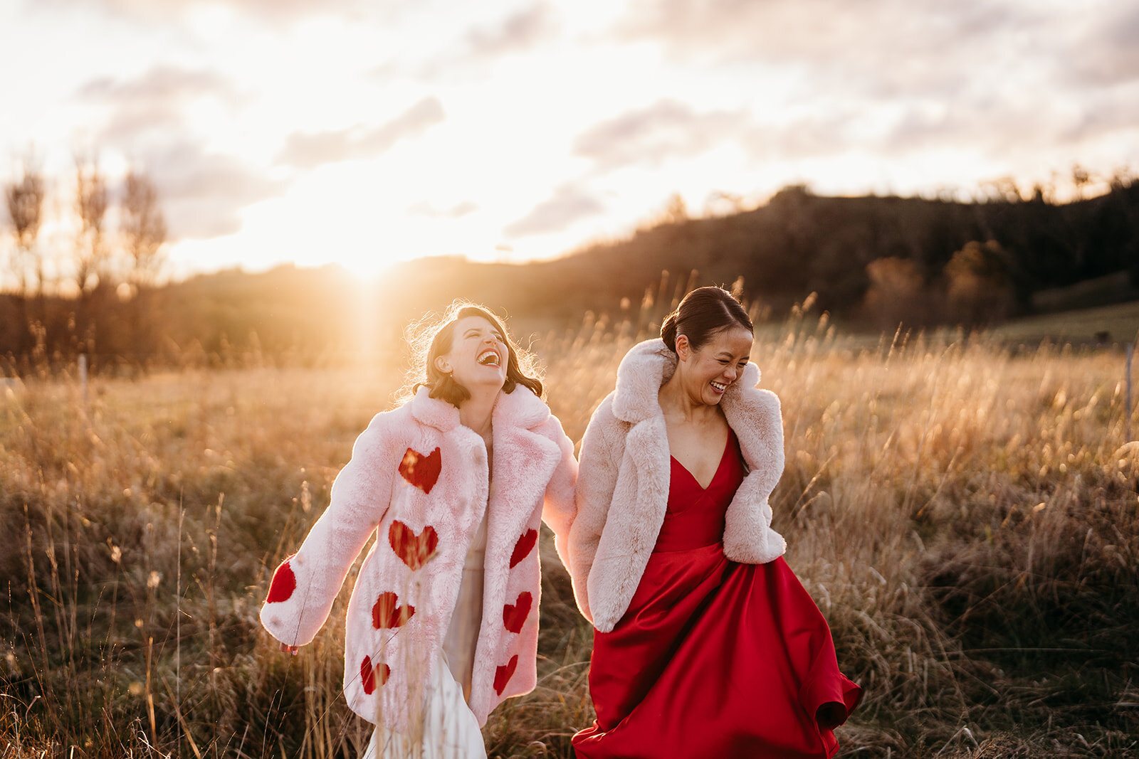Two brides in a golden field at sunset, one in a pink heart coat